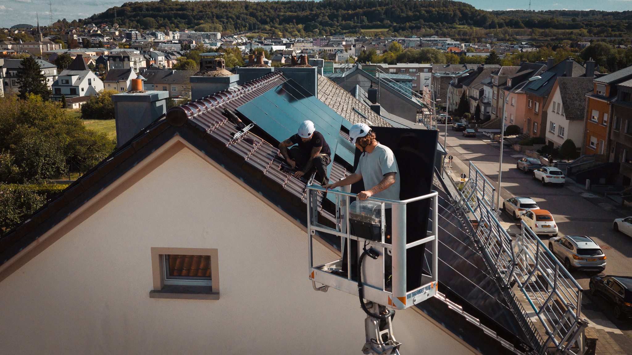 Two technicians install photovoltaic panels on a tiled roof using an aerial work platform.